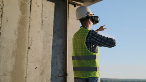 Engineer-Builder-visualizes-the-building-plan-standing-on-the-roof-of-the-building-at-sunset-stands-in-VR-glasses-and-moves-his-hands-using-the-interface-of-the-future.-Futuristic-engineer-of-the-future.-The-view-from-the-back.